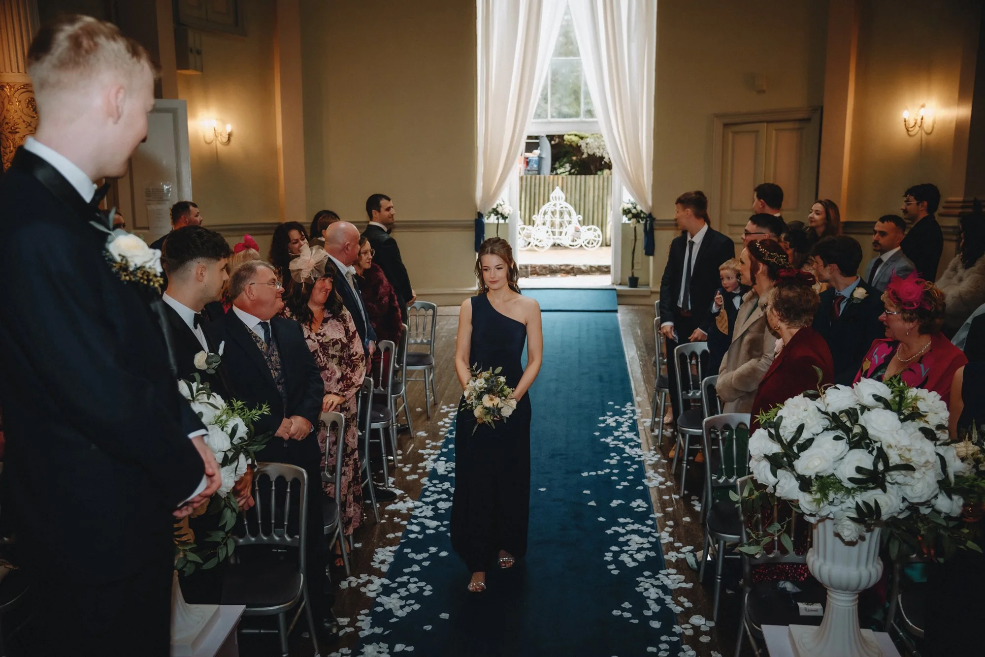 A woman in a navy blue dress walking down the aisle of a wedding ceremony, holding a bouquet, with guests seated on either side, petals scattered on the aisle, and an open door with curtains and a decorative carriage outside in the background.