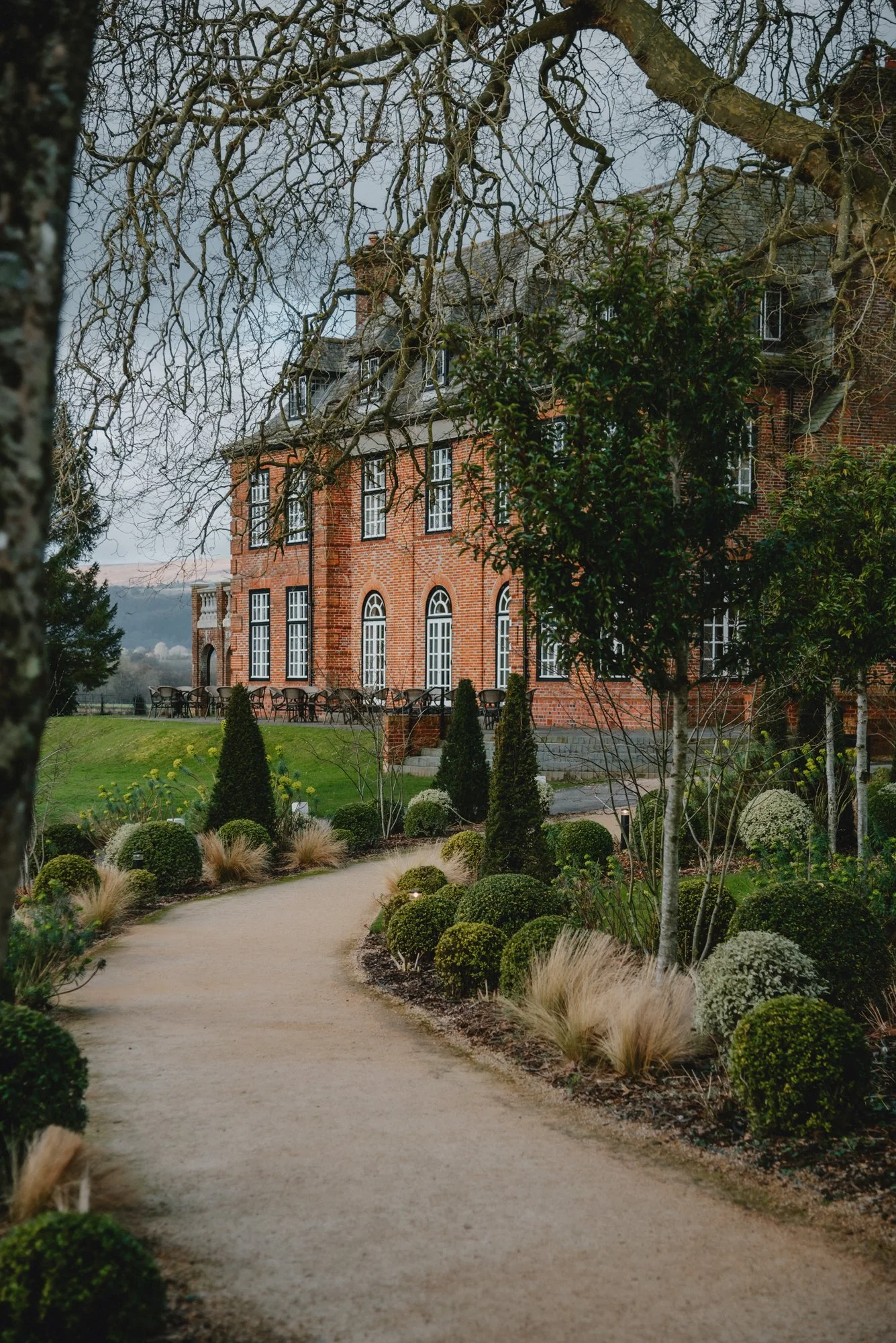 A brick mansion surrounded by a garden with trimmed bushes, trees, and a gravel pathway.