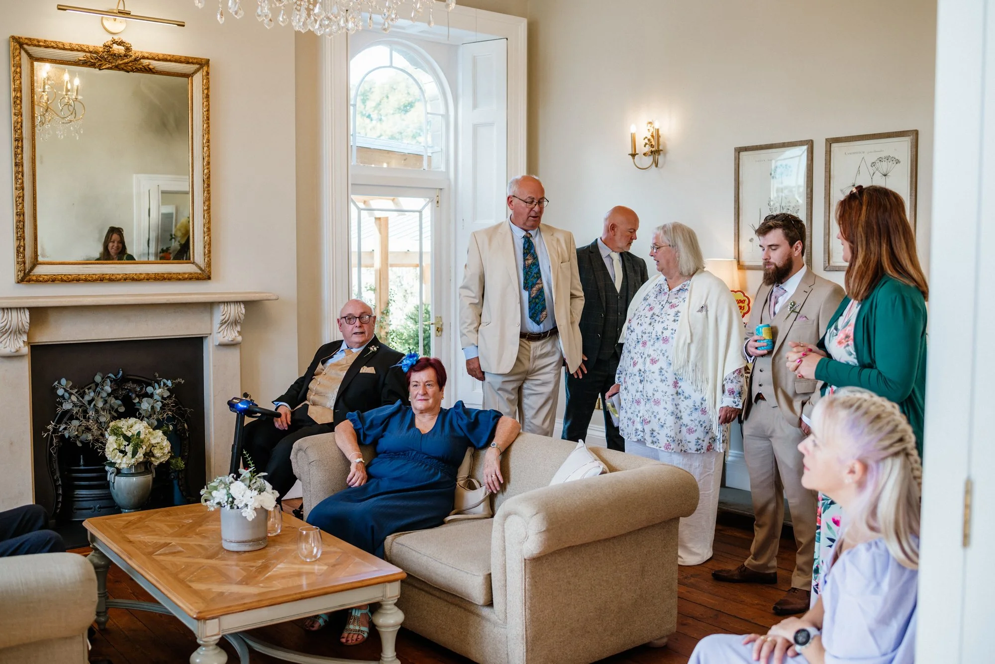 A group of people gathered in a bright living room with vintage decor, including a fireplace with a decorative flower arrangement, a large mirror, and framed botanical prints, engaging in conversation.