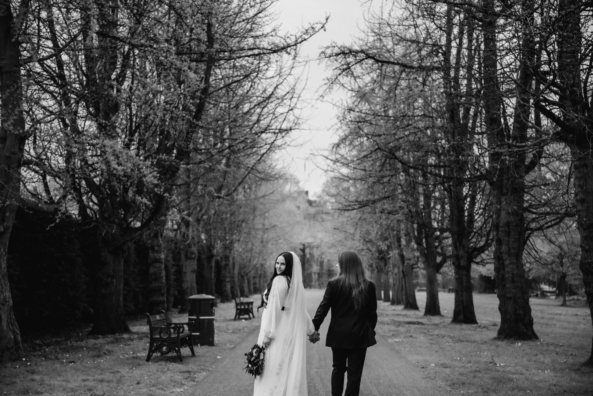 Black and white photo of a bride and groom walking hand in hand in a park lined with tall, bare trees.