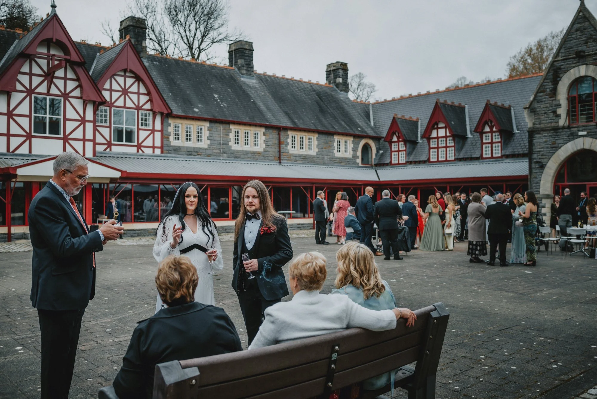 People gathered at an outdoor wedding or event in front of a large historic building with gray stone walls, red window frames, and Gothic-style architecture, during cloudy weather.