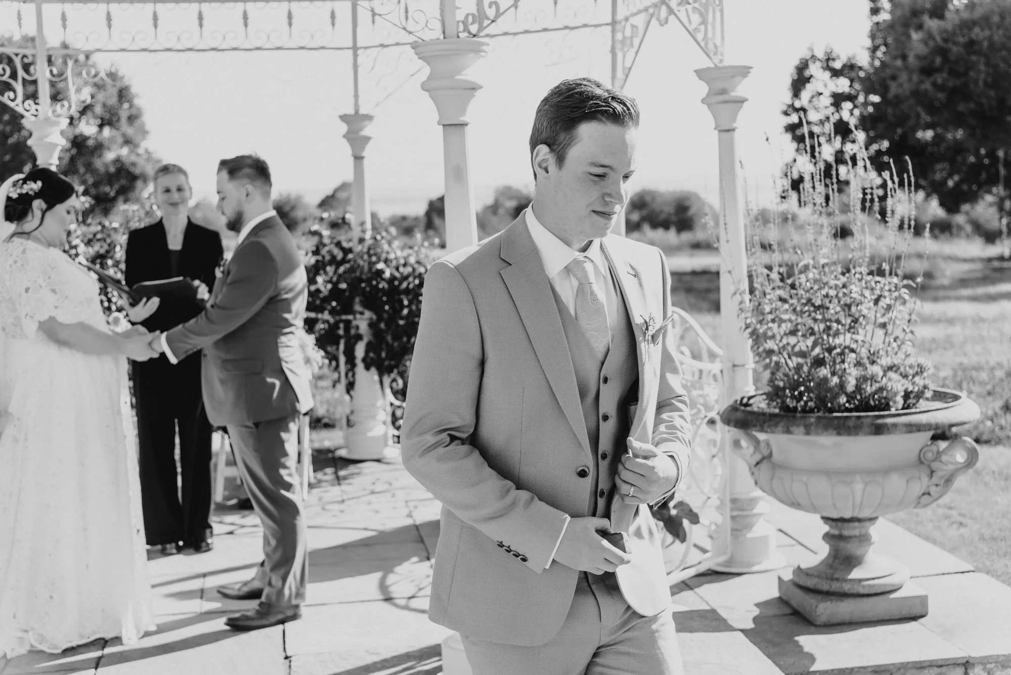 A groom in a light-colored suit, standing outdoors during a wedding ceremony, appears to be emotional as he looks down with his eyes closed, while the bride and officiant are visible in the background exchanging vows.
