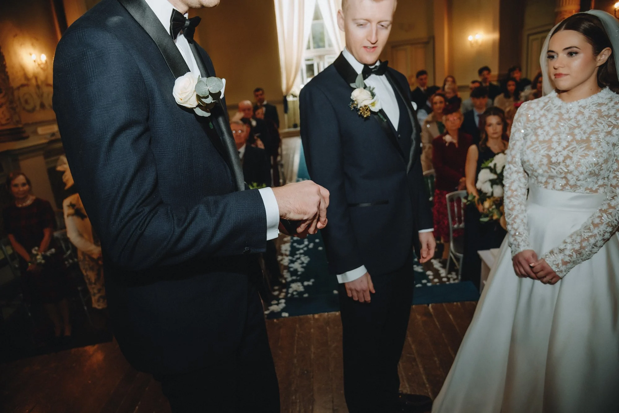 A wedding ceremony with two grooms and a bride standing in front of guests, with the grooms holding hands and the bride standing with her hands clasped.