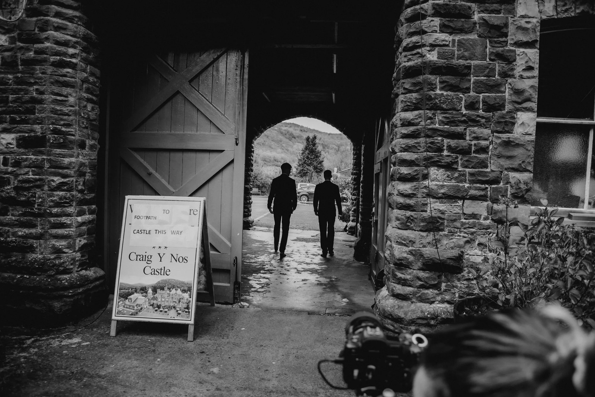 Two men in suits walking out of a stone archway, with a signboard for Craig Y Nos Castle on the left and a window on the right, in black and white.