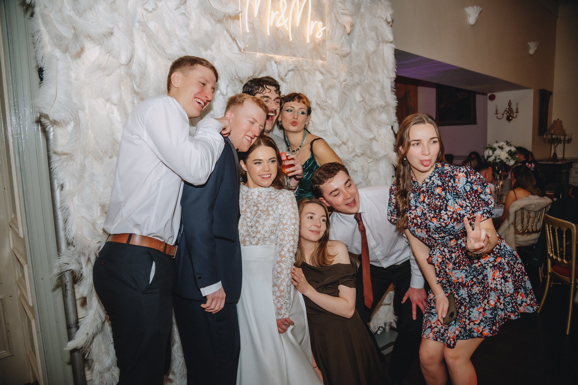 Group of young people at a wedding celebration, posing and making playful gestures in front of a feathered backdrop with a neon sign that reads 'Mr & Mrs'.