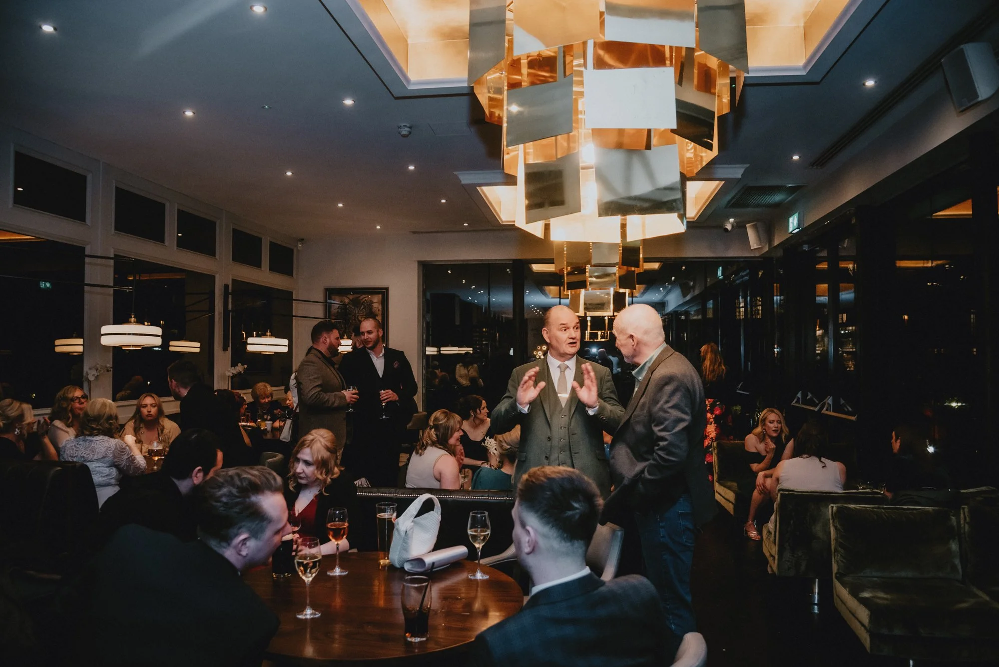 People socializing at a formal dinner event in an upscale restaurant or banquet hall, with a modern chandelier overhead.