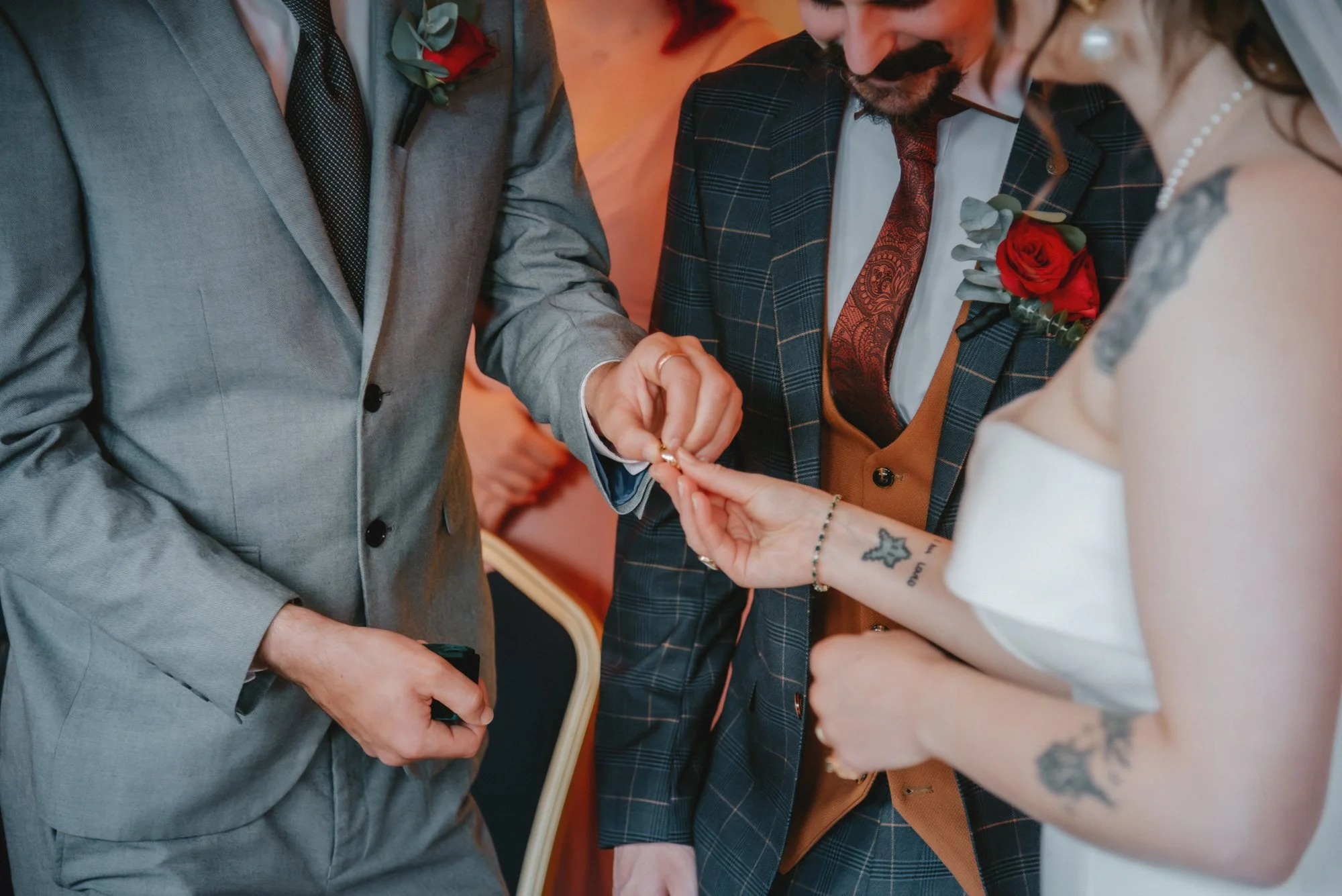 A wedding ceremony where a person is placing a ring on the bride's finger, with the groom and bride dressed in formal attire with floral boutonnieres and tattoos visible on the bride's arm.