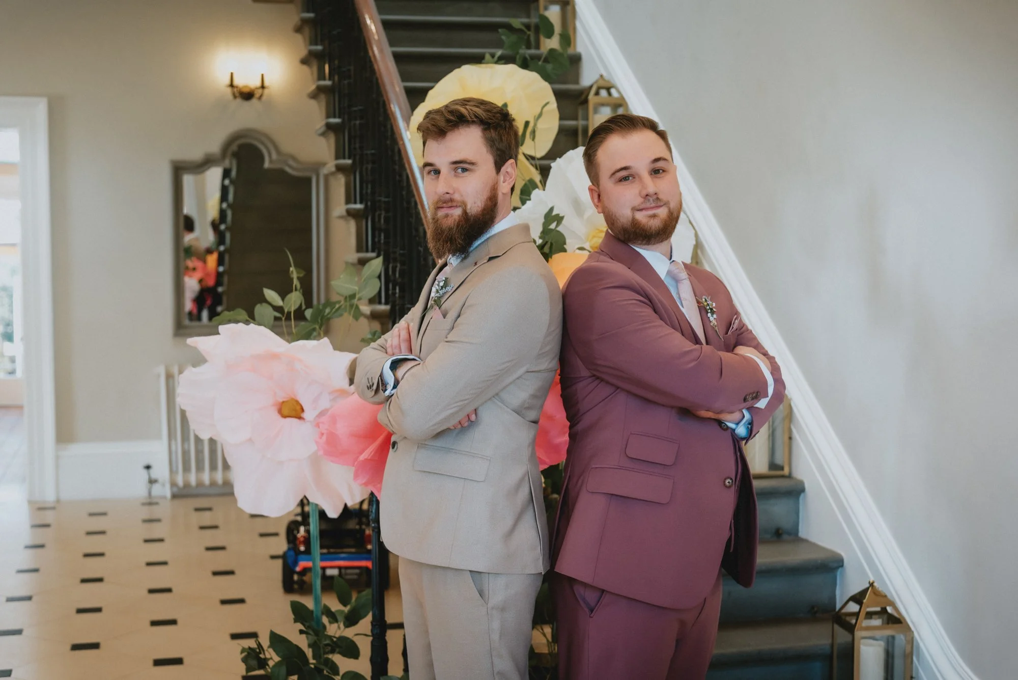 Two men in suits standing back-to-back with arms crossed, posing in front of a floral arrangement inside a building with staircase and wall decor.