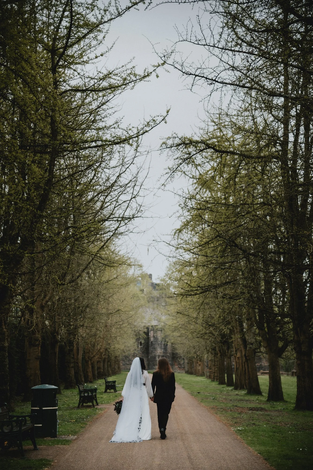 A bride wearing a white wedding dress and veil holding a bouquet walks hand-in-hand with a woman in a black suit down a tree-lined park path.