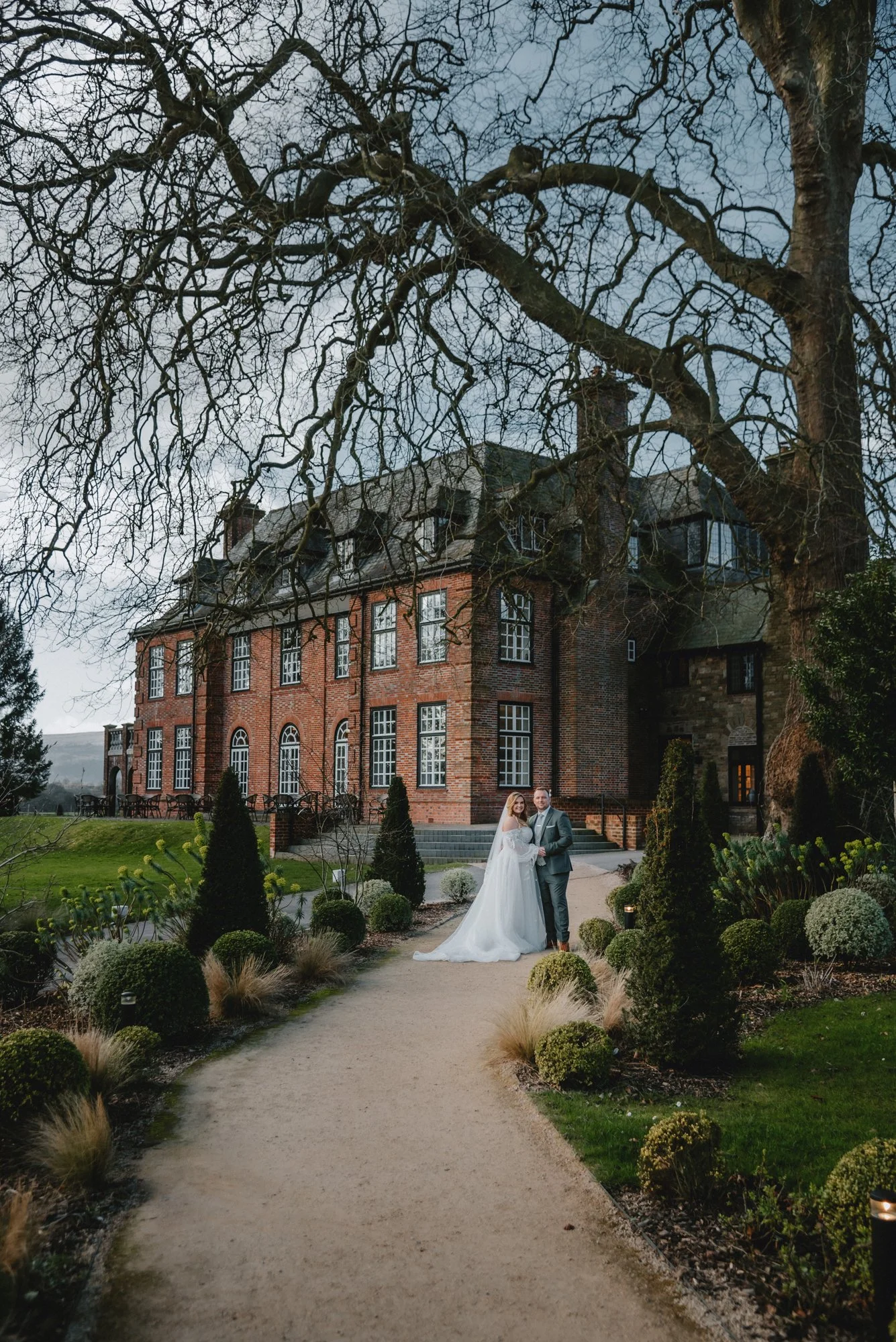 A bride and groom standing on a garden path in front of a large brick mansion during their wedding, with a leafless tree overhead and landscaped bushes surrounding them.