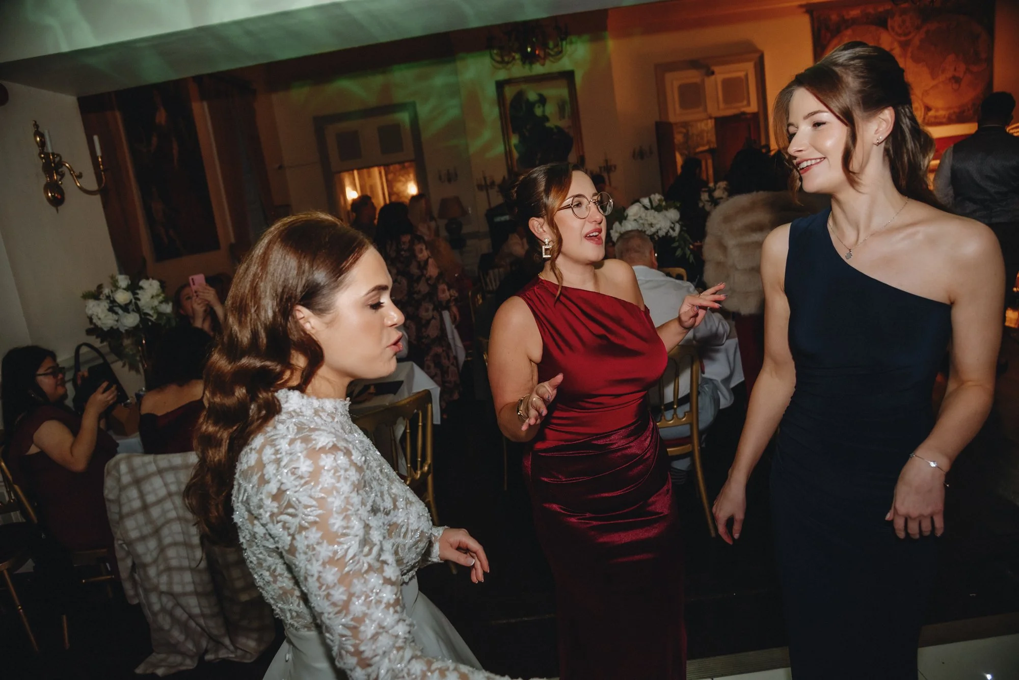 Three women dancing and talking at a formal indoor event, with tables and floral arrangements in the background.