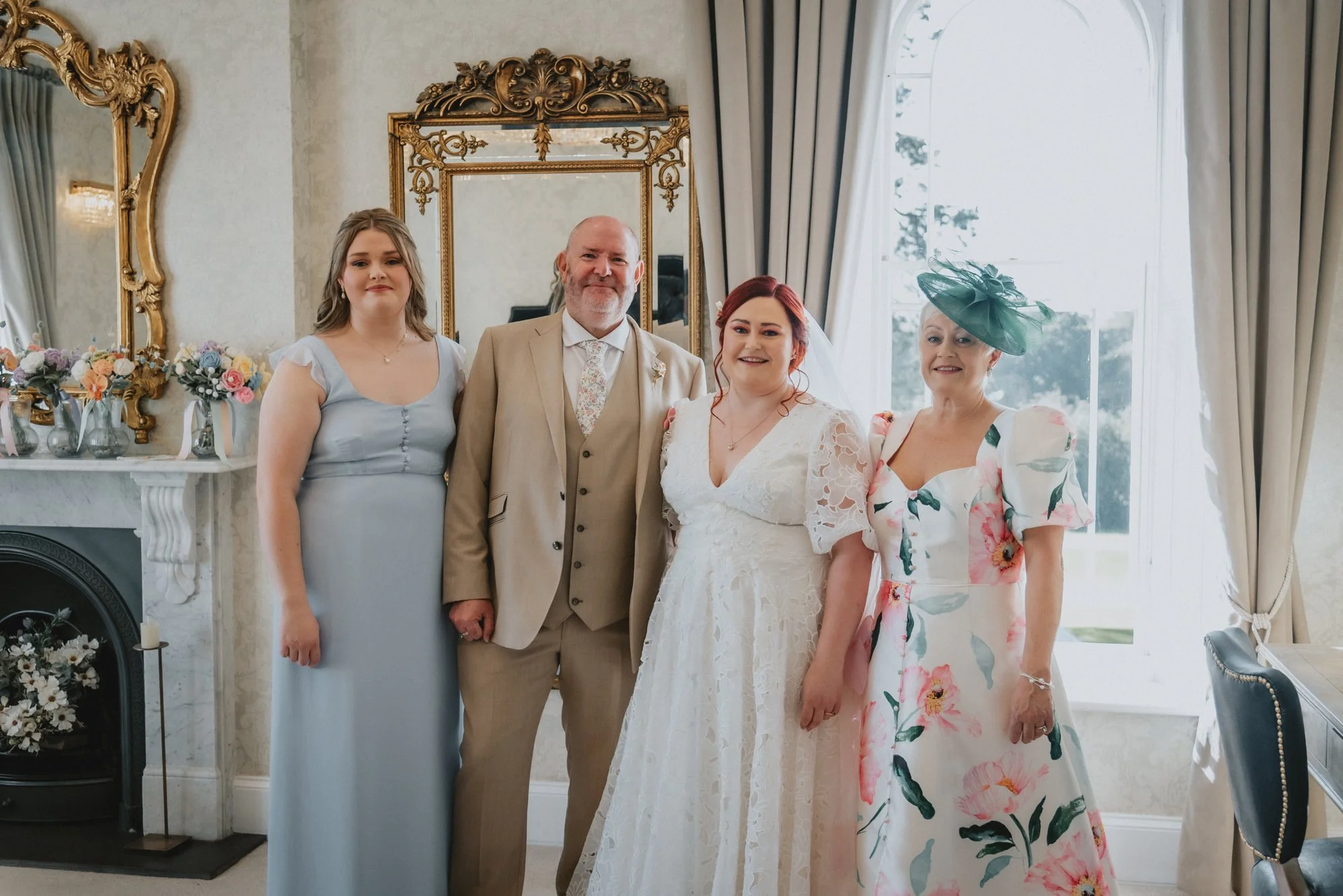 Group of four people, including a bride in a white lace wedding dress, standing inside a decorated room with large windows, curtains, floral arrangements, and ornate mirrors.