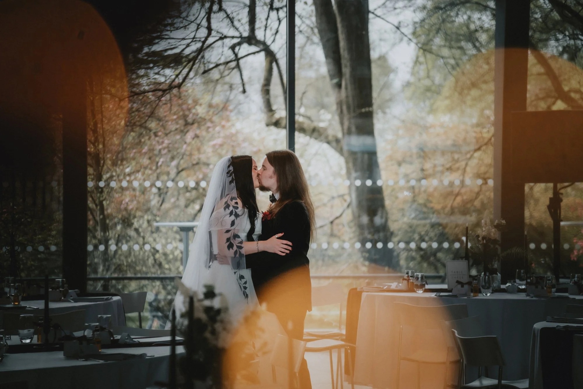 Two women share a kiss at a wedding reception indoors with large windows showing autumn trees outside, decorated tables, and floral arrangements.