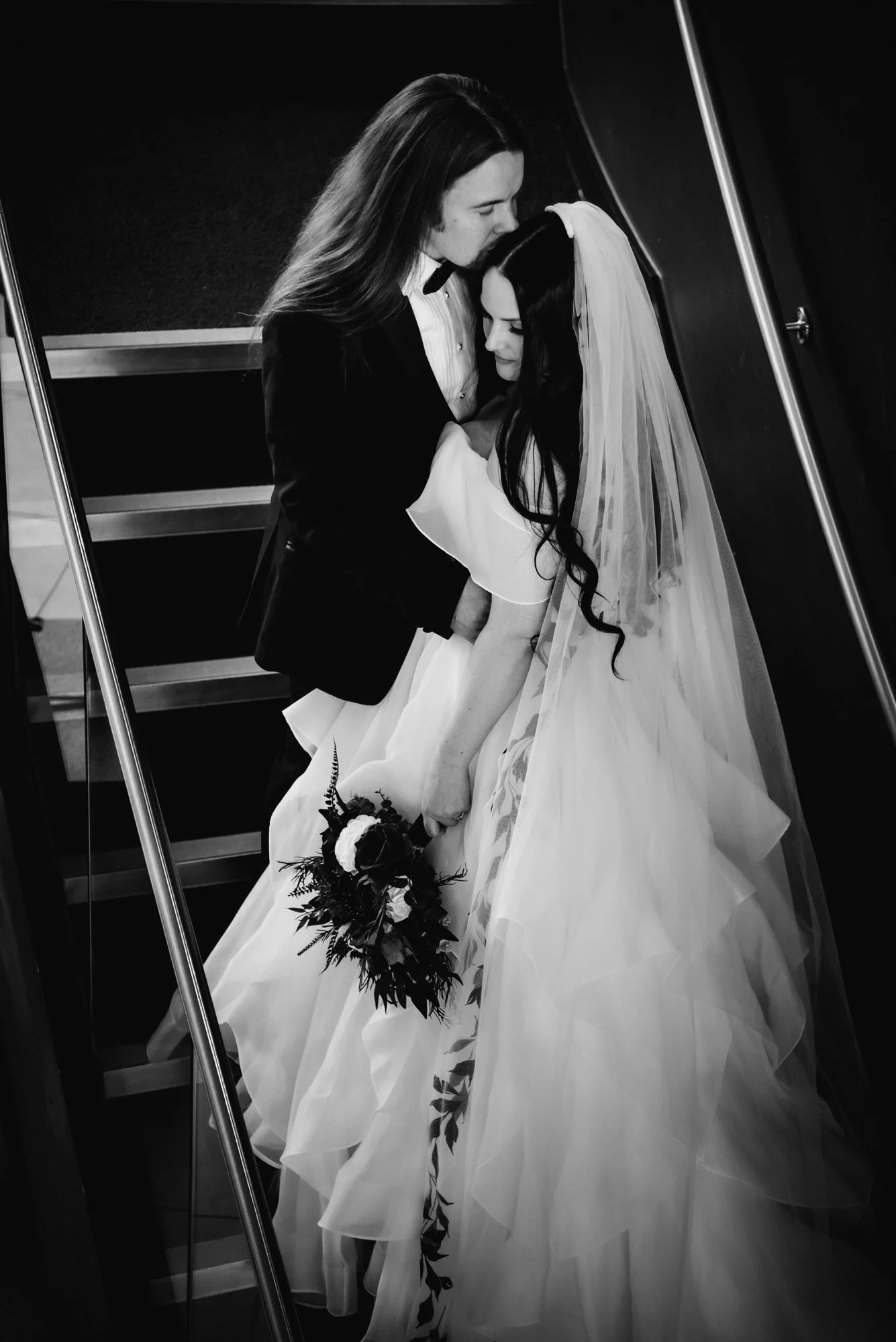 A black and white photo of a bride and groom embracing, with the groom kissing the bride's forehead. The bride is holding a bouquet, and her wedding dress has a long veil.