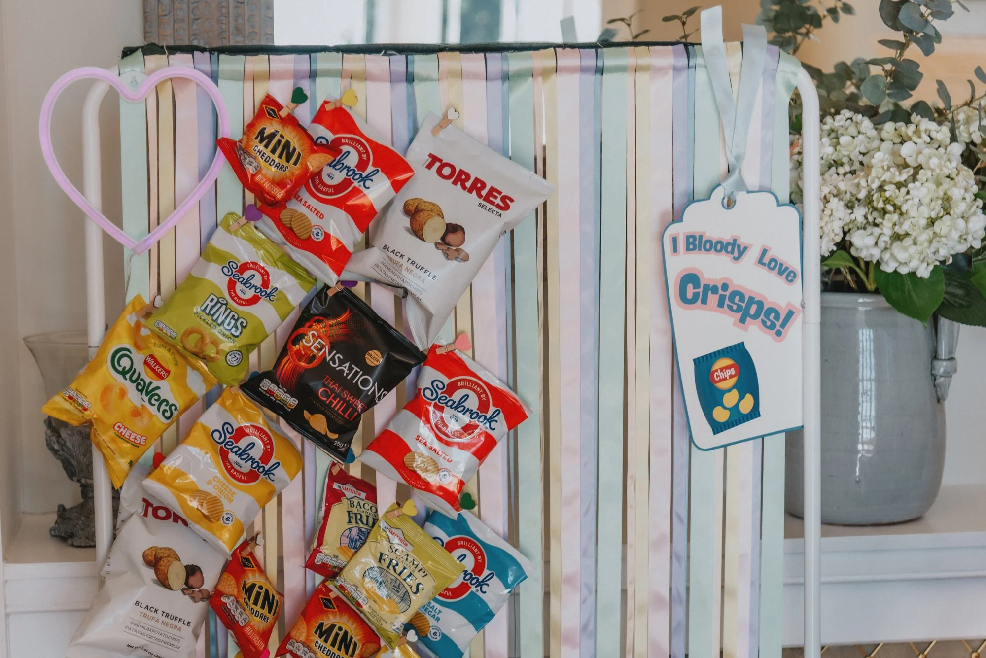 Decorative display of various snack bags hanging on a gift card holder, with a large floral arrangement in a gray vase in the background. The snack bags include popcorn, chips, and other savory snacks, with a sign that reads "I Bloody Love Crisps!" f