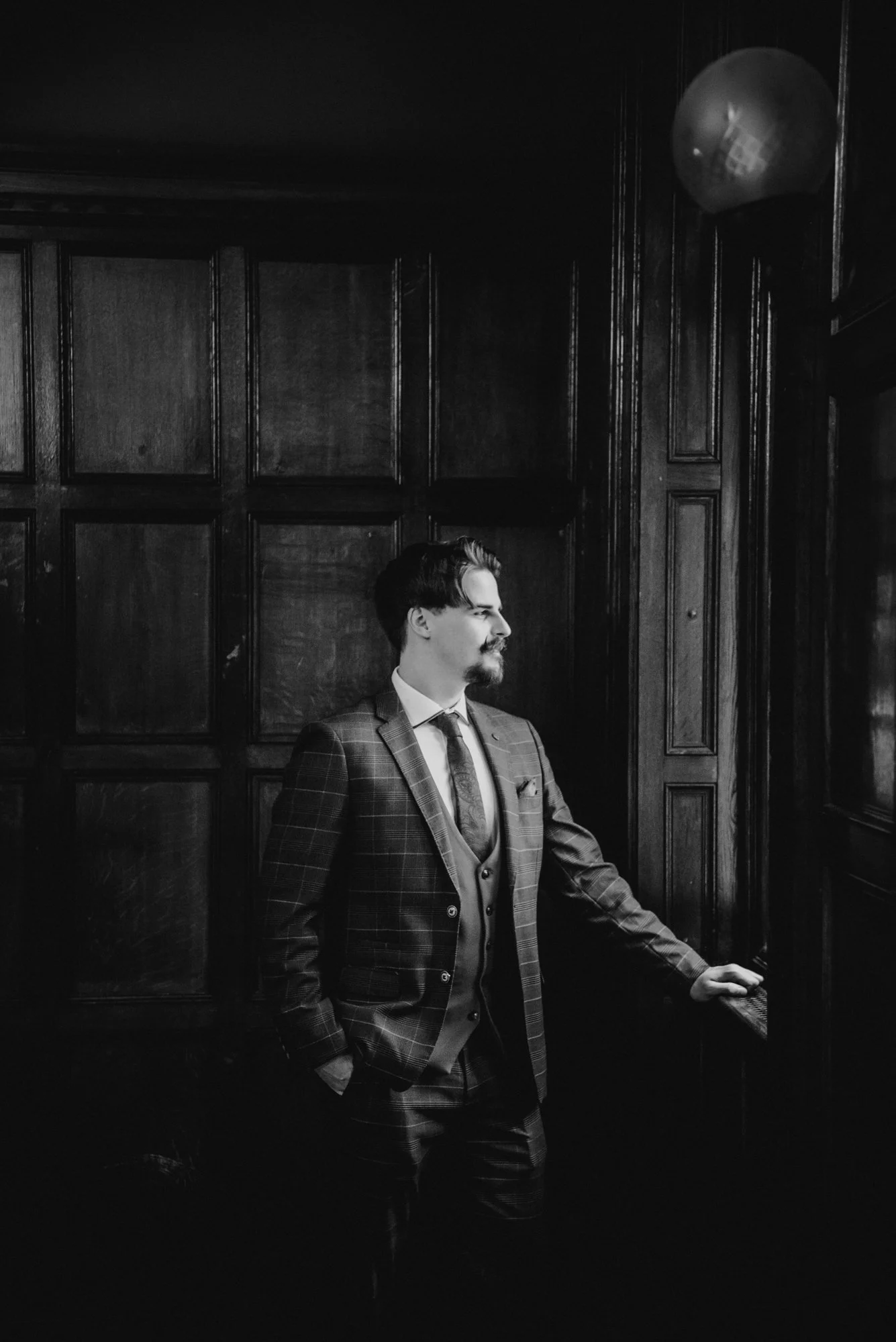 A man in a suit standing by a window in a wood-paneled room, looking outside.
