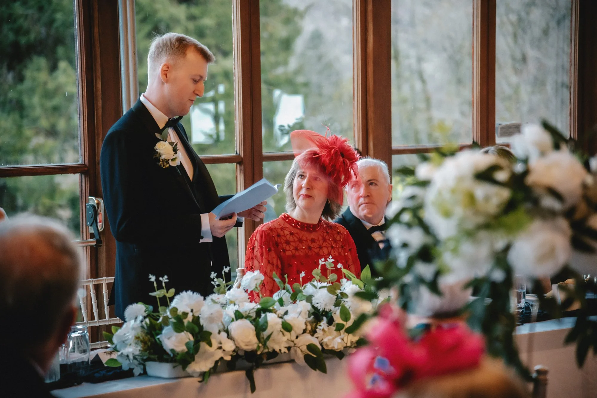 A young man in a tuxedo giving a speech at a wedding reception, standing beside two seated guests, a woman in a red dress and a man in a black tuxedo, with floral decorations on the table and large windows in the background.