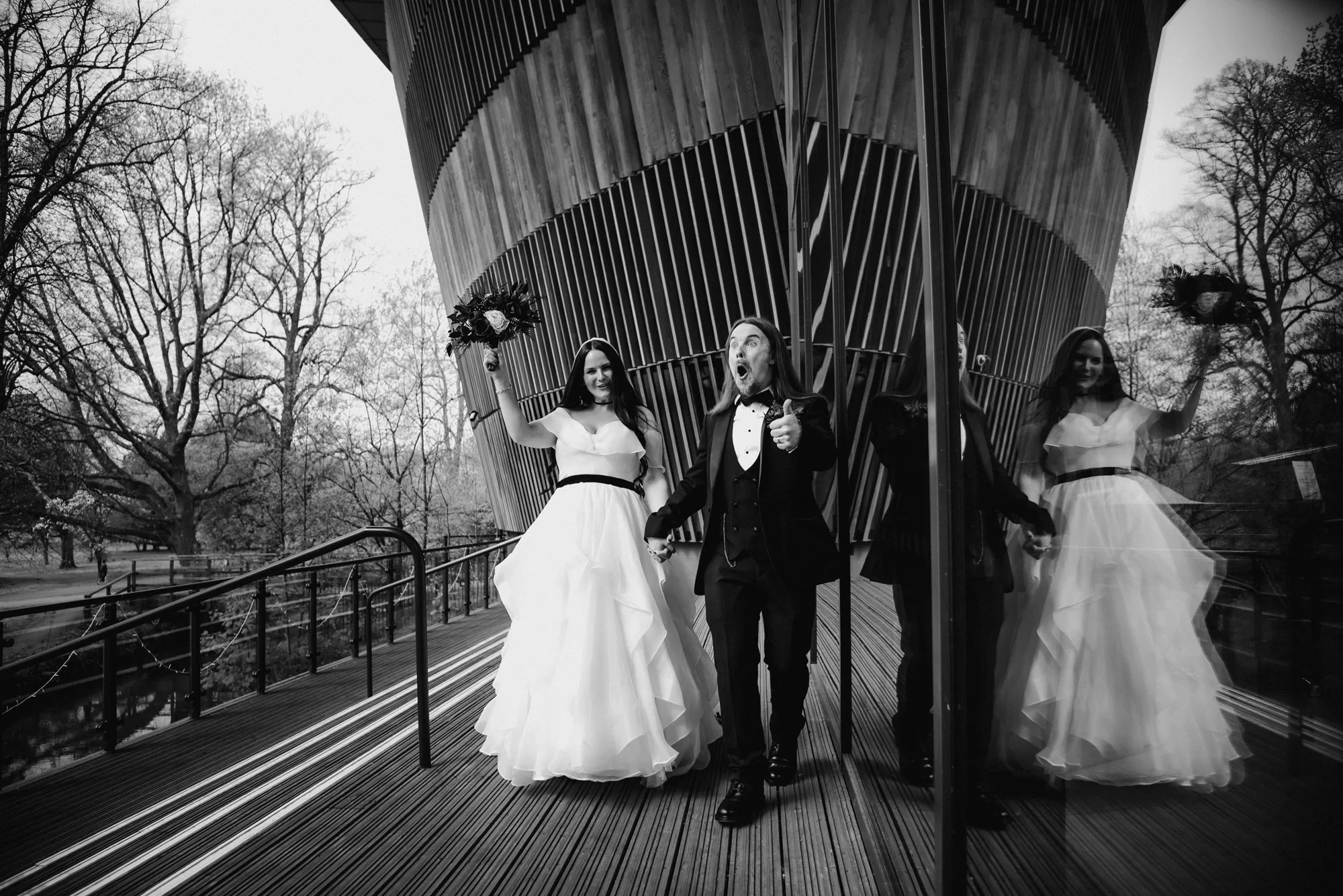 Black and white photo of three people standing outside in front of a modern building with glass walls. Two women in wedding dresses are on either side of a man in a tuxedo, who is giving a thumbs up. The woman on the left is holding a bouquet and smi