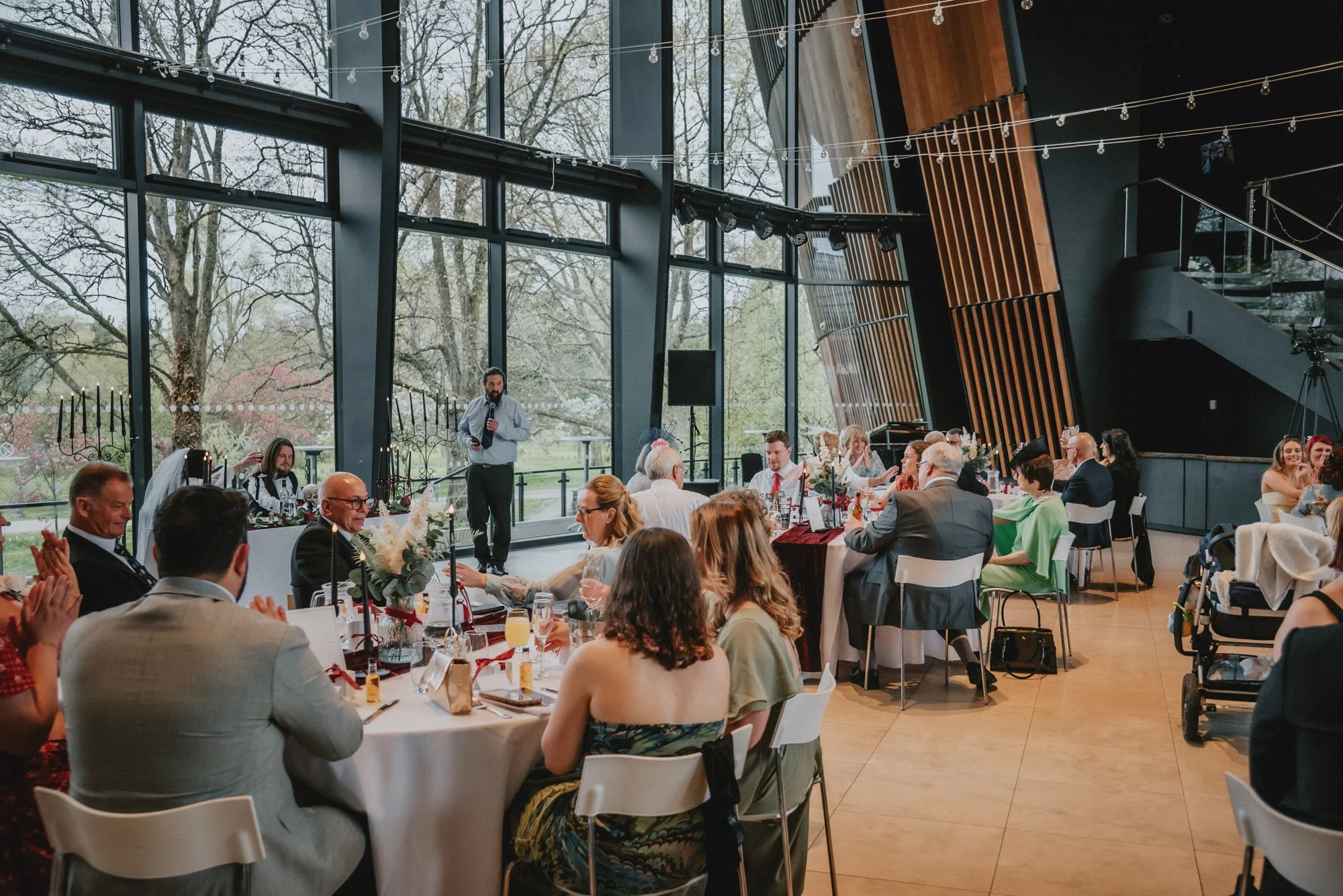 Indoor wedding reception with guests seated at round tables, a man giving a speech at a microphone, and large windows showing trees outside, decorated with string lights.