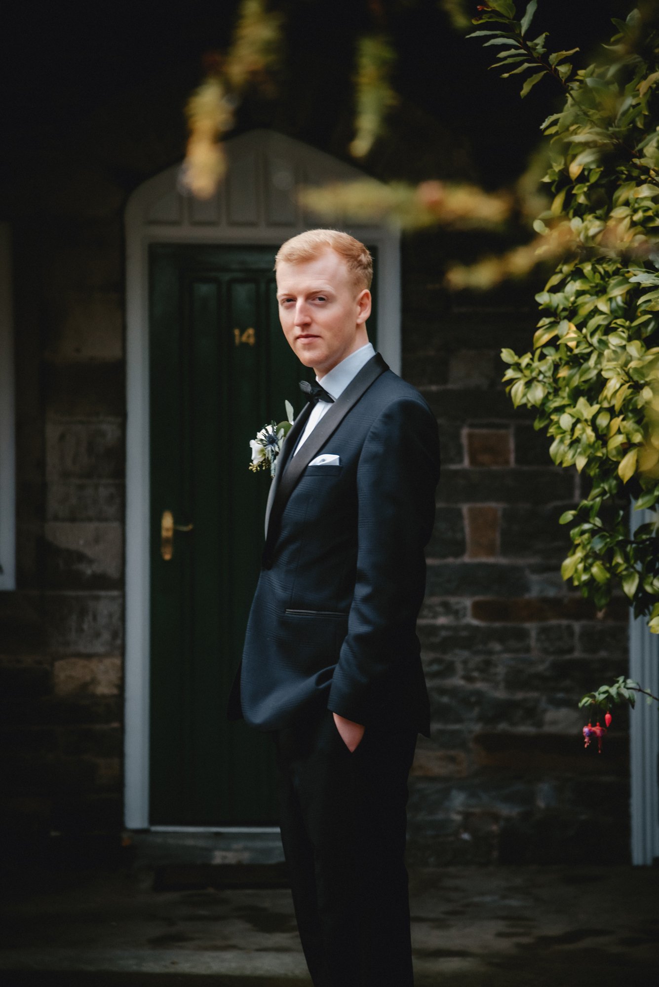 A young man in a black tuxedo with a bow tie, standing outdoors in front of house number 14, posing with one hand in his pocket, surrounded by greenery.