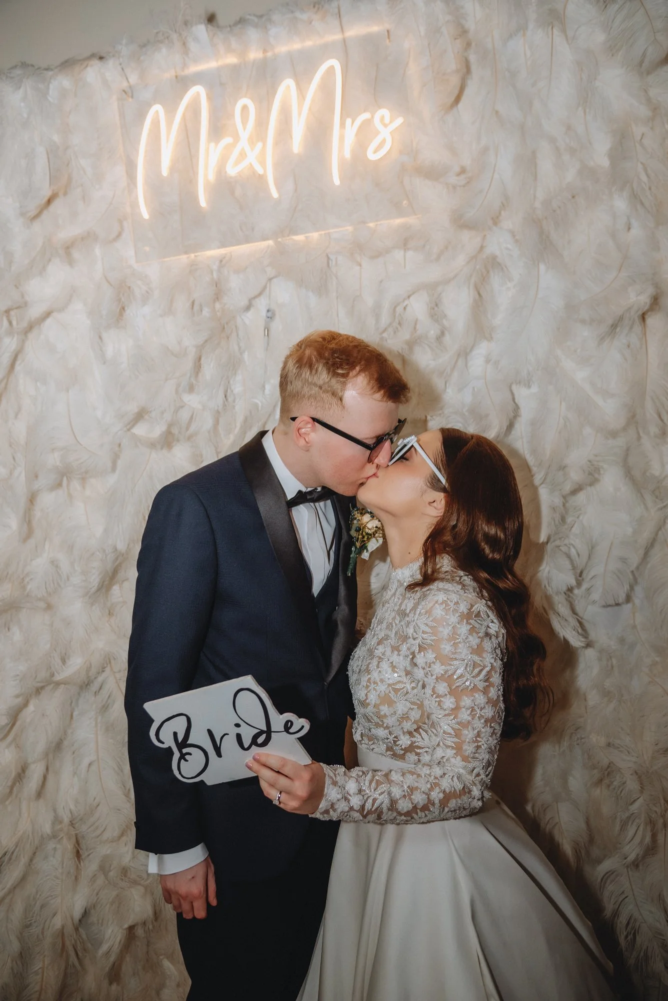 Couple in wedding attire sharing a kiss at their wedding reception, with a neon sign reading 'Mr & Mrs' and a woman holding a 'Bride' sign.