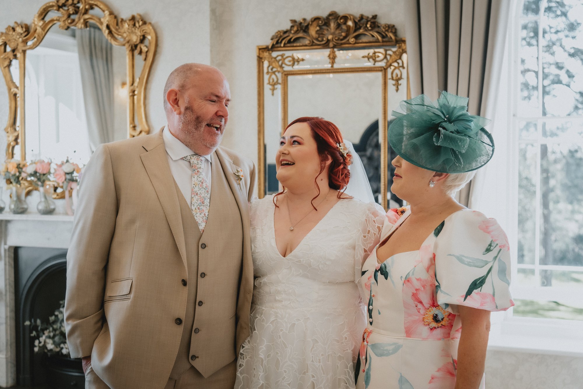 A wedding scene with three people: a man in a beige suit, a woman in a white wedding dress, and another woman in a floral dress with a large green hat, standing indoors with mirrors and floral decorations.
