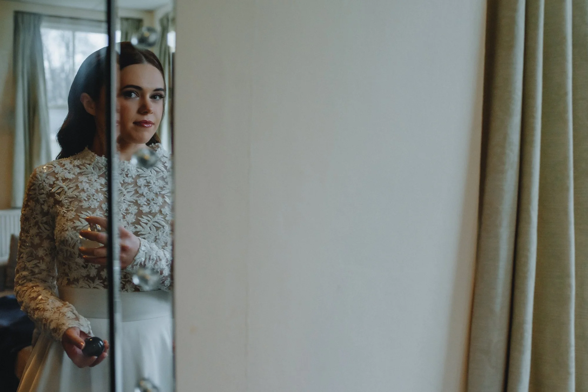 A woman dressed in a wedding gown with lace details, looking at her reflection in a mirror.