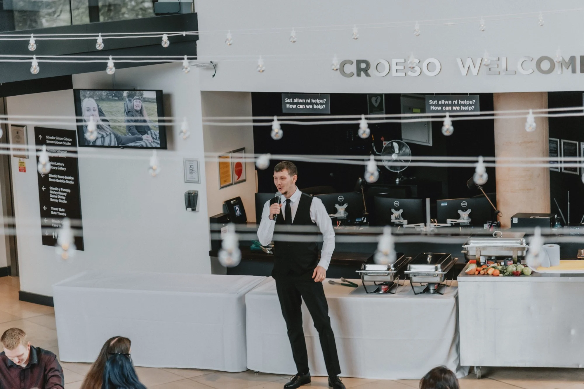 A man in a formal vest and tie, holding a microphone, speaking in front of an audience in a modern event space with string lights hanging above and a buffet table to the right, with several chairs and people seated in the foreground.