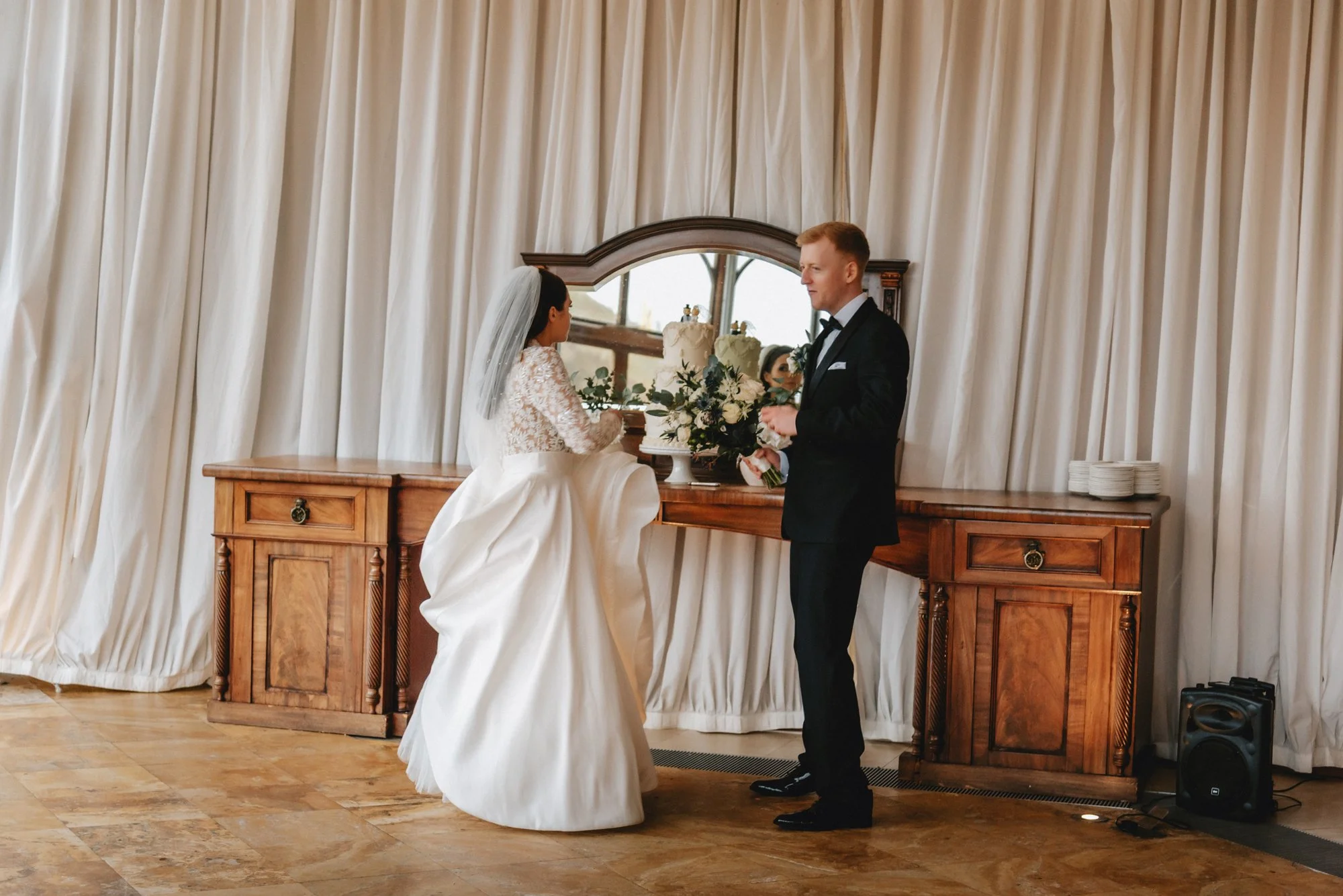 Bride and groom standing in front of a cake at their wedding, with a mirror and curtains behind them.