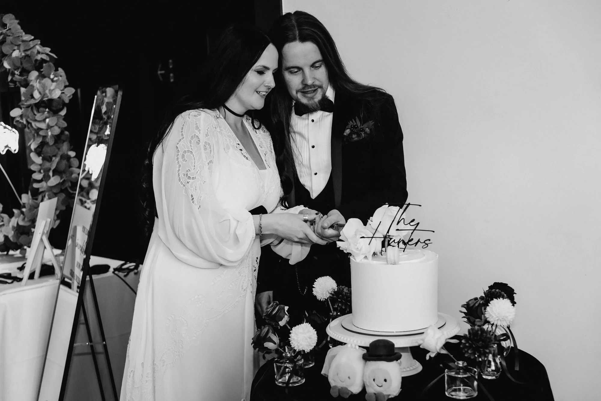A couple in wedding attire cutting a wedding cake at a celebration, with flowers and decorative items on the table.