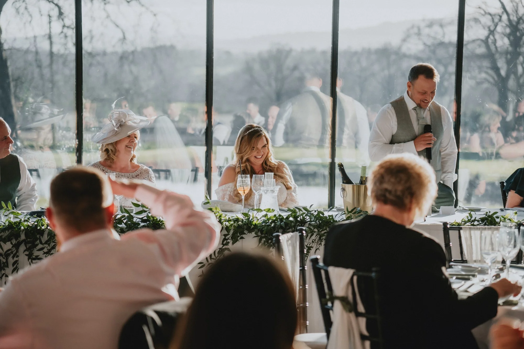 Wedding reception with people sitting at a long table, a man giving a speech with a microphone, women dressed in formal attire, and a window view of an outdoor landscape with trees.