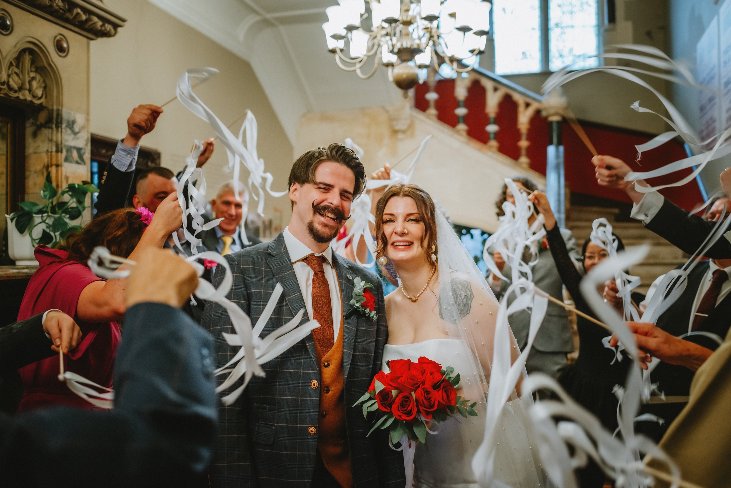 Happy couple, the groom in a navy suit and the bride in a white dress, celebrating with arms raised and smiling in front of a gray metal wall, the bride holding a bouquet.