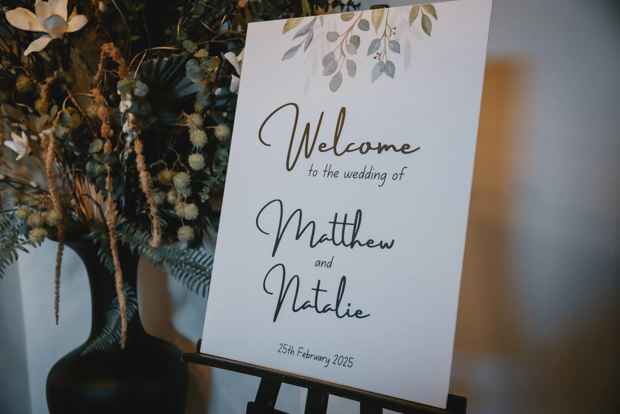 Wedding welcome sign with green foliage design at the top, featuring the text 'Welcome to the wedding of Matthew and Natalie' and the date '25th February 2025', placed on a black stand next to a floral arrangement in a black vase.