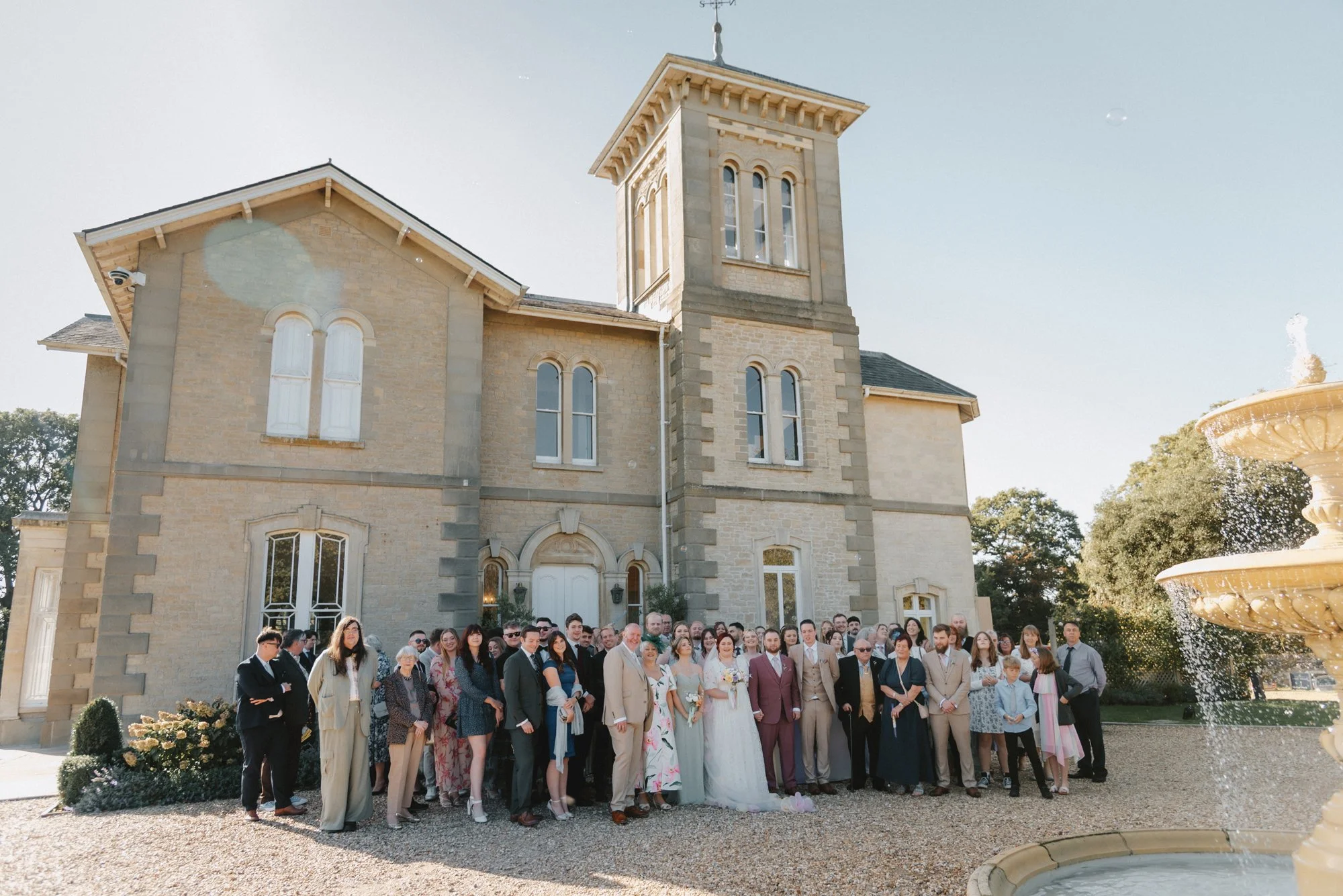 A large group of people gathered in front of a historic stone mansion during a wedding celebration on a sunny day, with a fountain visible on the right