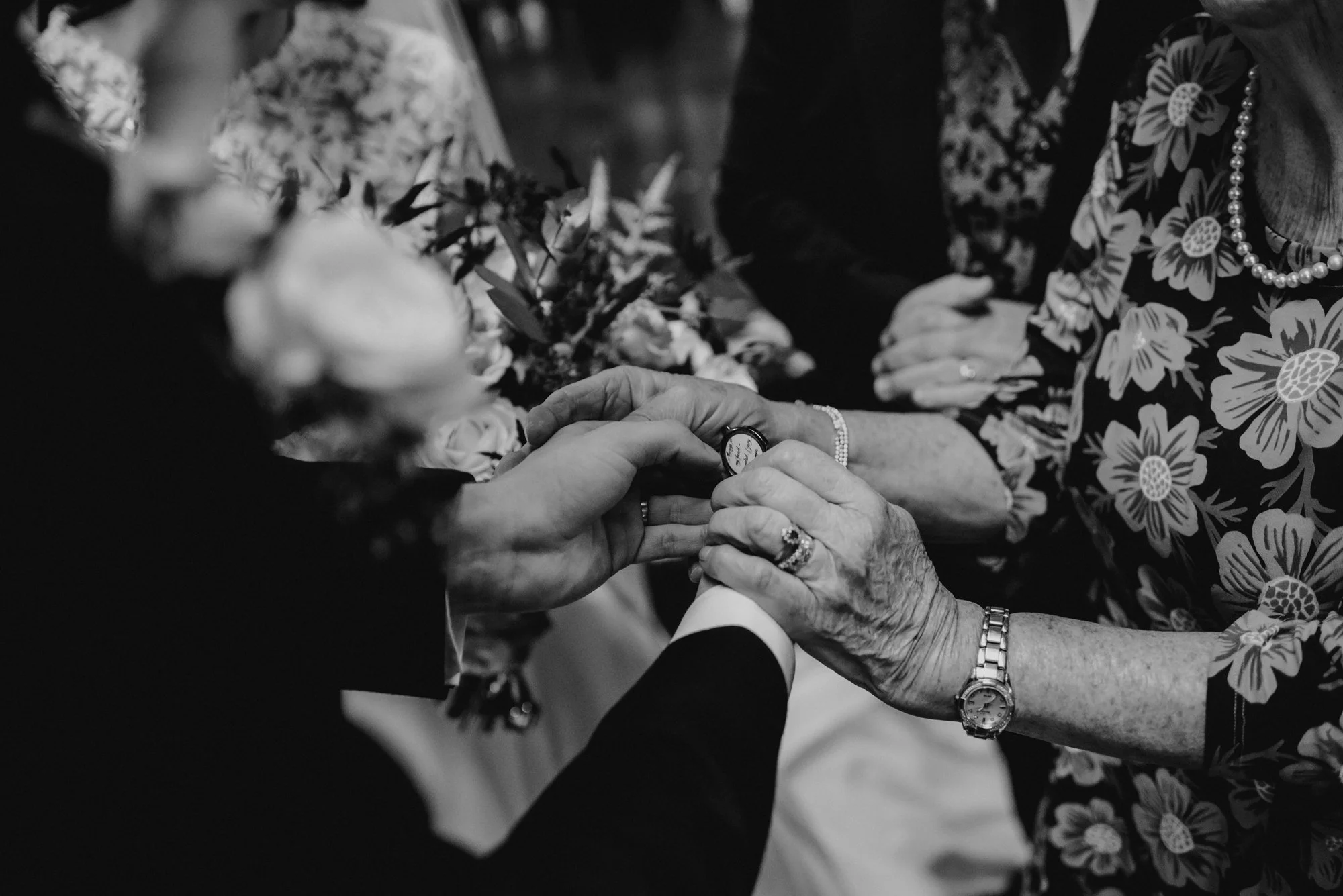 A wedding ceremony where a person is placing a ring on another person's finger, with another woman holding their hand and wearing a floral dress, pearl necklace, and watch, amidst flowers and formal attire.