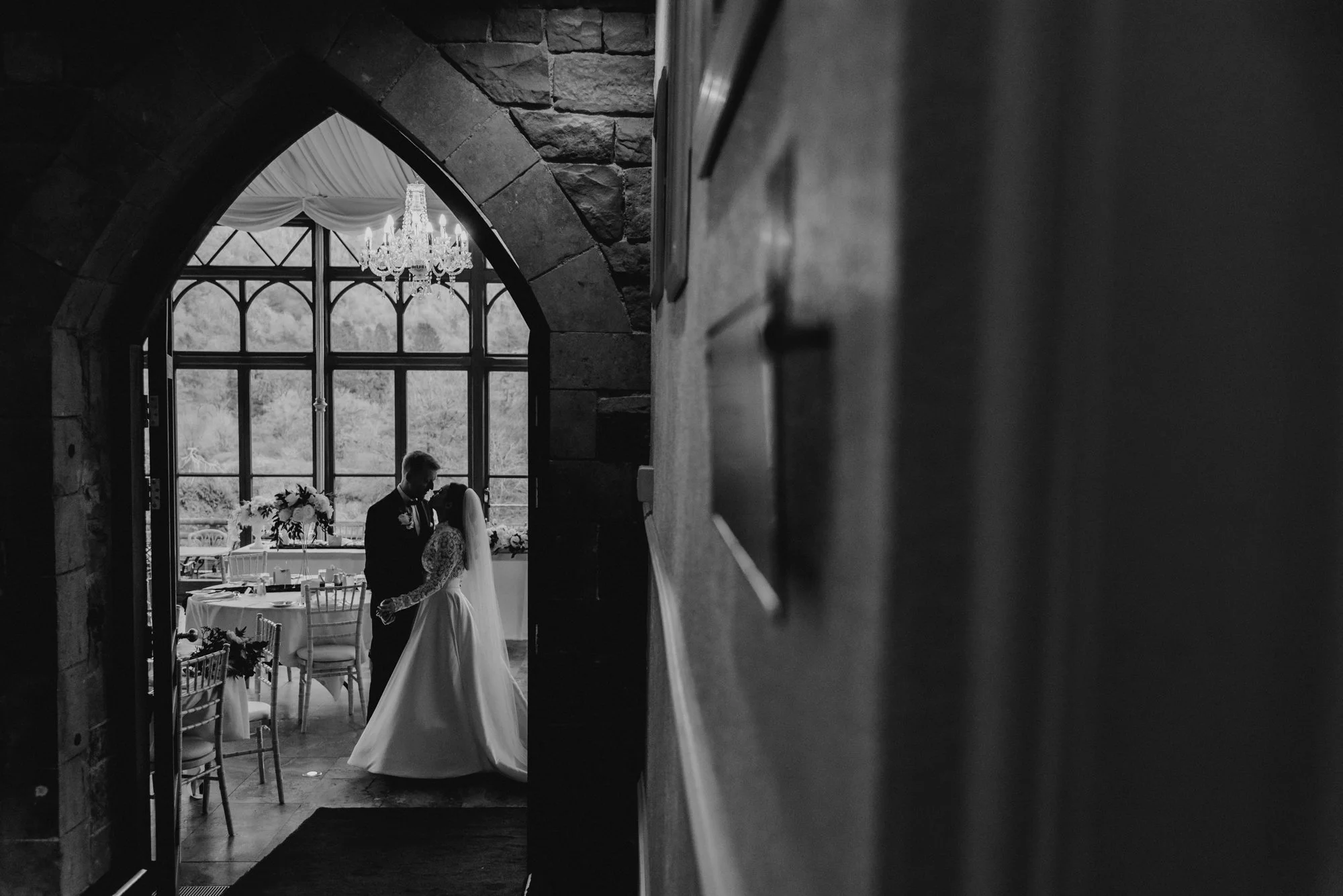 A black-and-white photo of a bride and groom dancing in a decorated wedding reception area viewed through an arched doorway, with a large window and chandelier in the background.