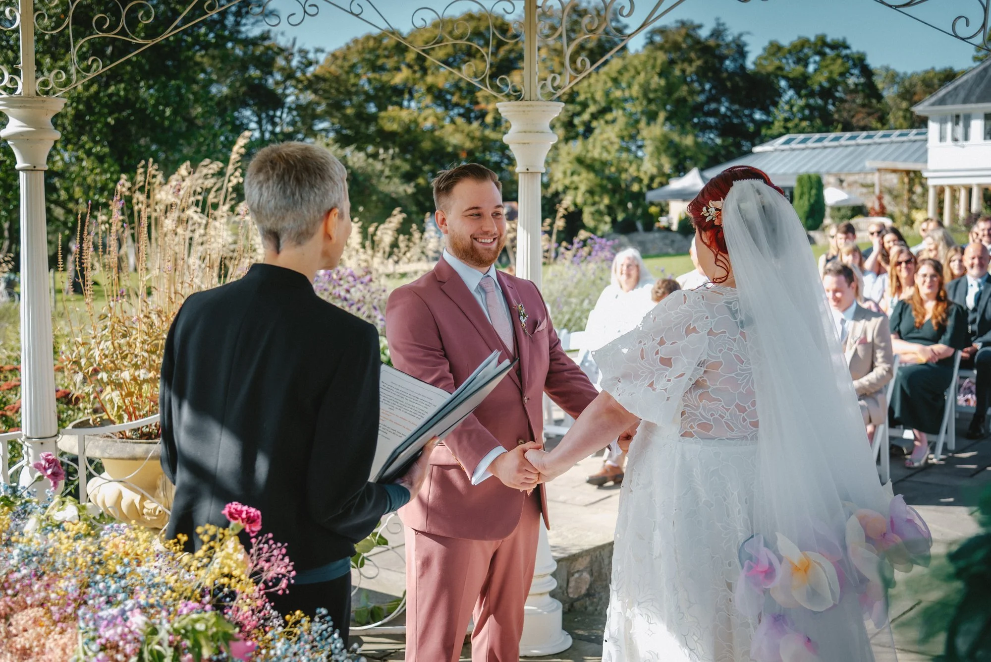 A wedding ceremony taking place outdoors with a smiling groom holding the bride's hands, officiant, and guests watching, under a decorative white gazebo with a garden in the background.