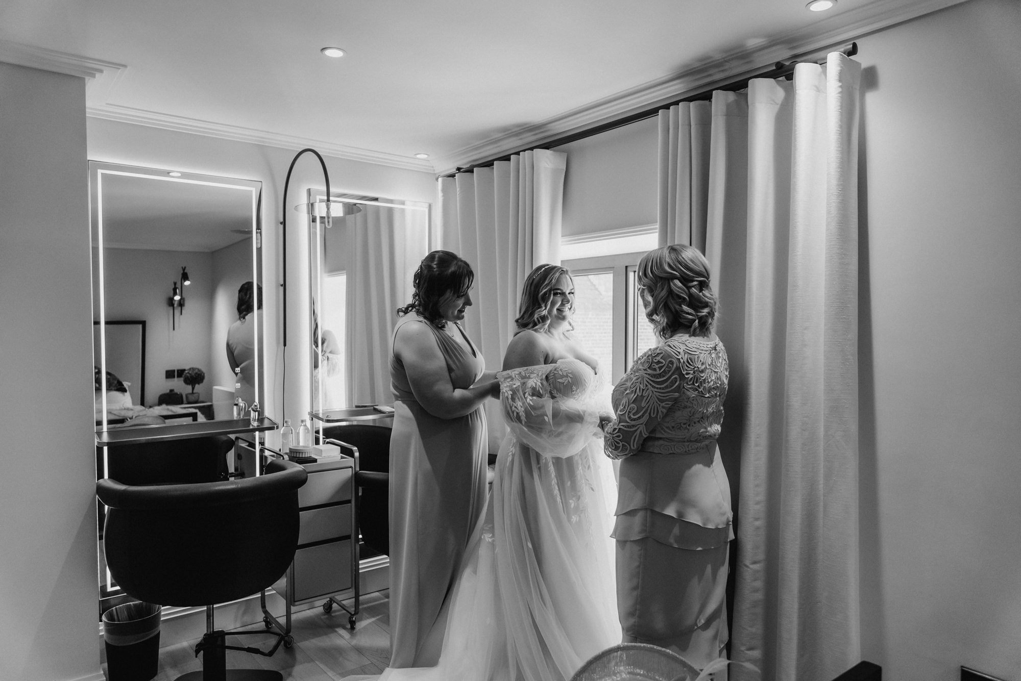 A bride getting ready with two women helping her, in a well-lit room with a window, curtains, and dressing tables.
