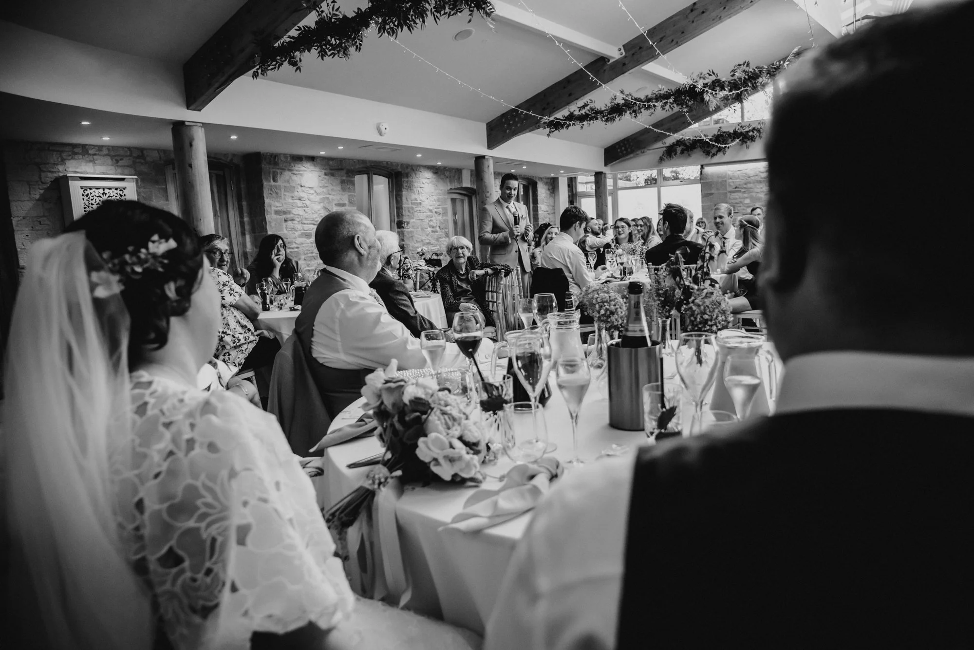 Black and white photo of a wedding reception with guests seated at tables and a man giving a speech while holding a microphone.