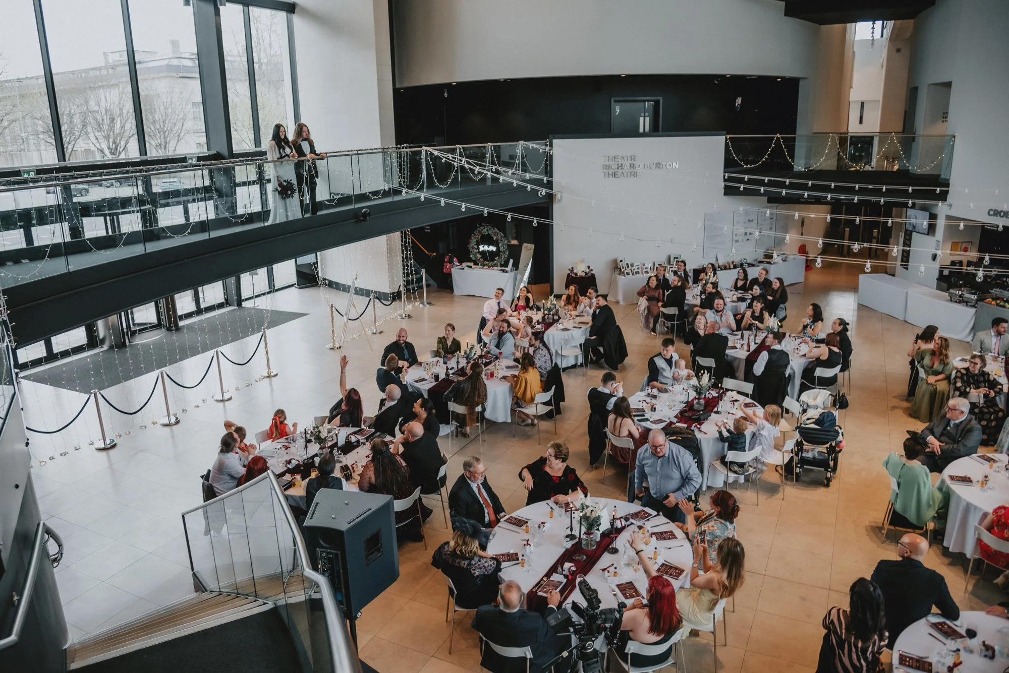 Guests seated at round tables during a formal event in a modern venue with large windows, holiday decorations, and string lights.