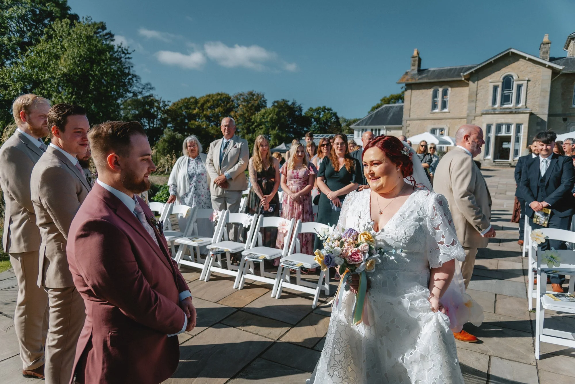 Bride smiling and holding a bouquet of flowers during her outdoor wedding ceremony, with guests seated and standing under a clear sky in the background.