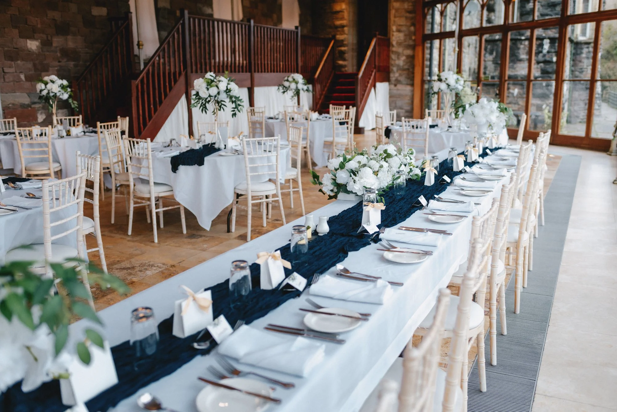 Elegant indoor wedding reception setup with long tables decorated with white tablecloths, navy blue runners, and white floral centerpieces; cream-colored chairs, large windows, and stone walls are visible.