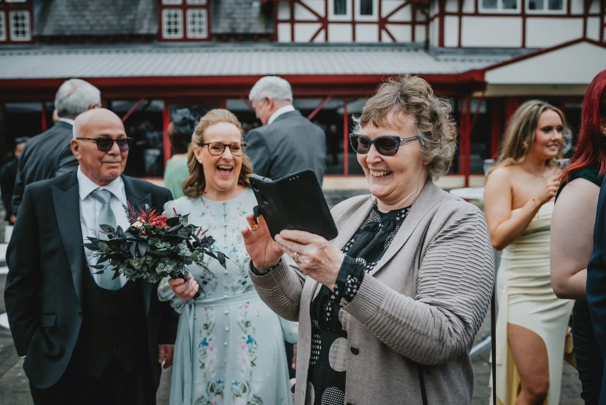 Group of people smiling and enjoying a social gathering, with one woman taking a photo on her phone, outside with a building in the background.