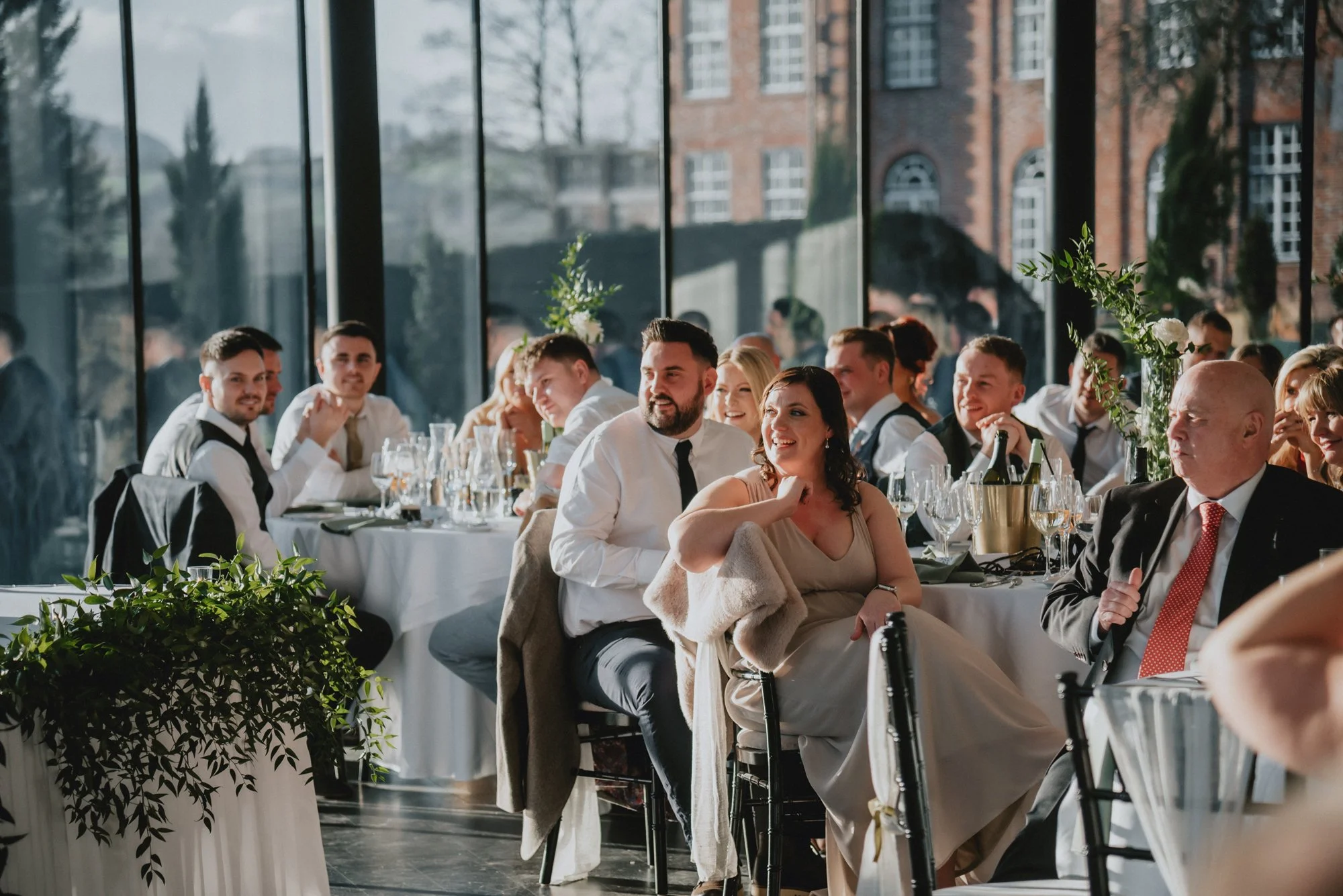 People seated at a formal event or wedding reception, smiling and engaging, inside a venue with large glass windows showing an outdoor scene with brick buildings and trees.