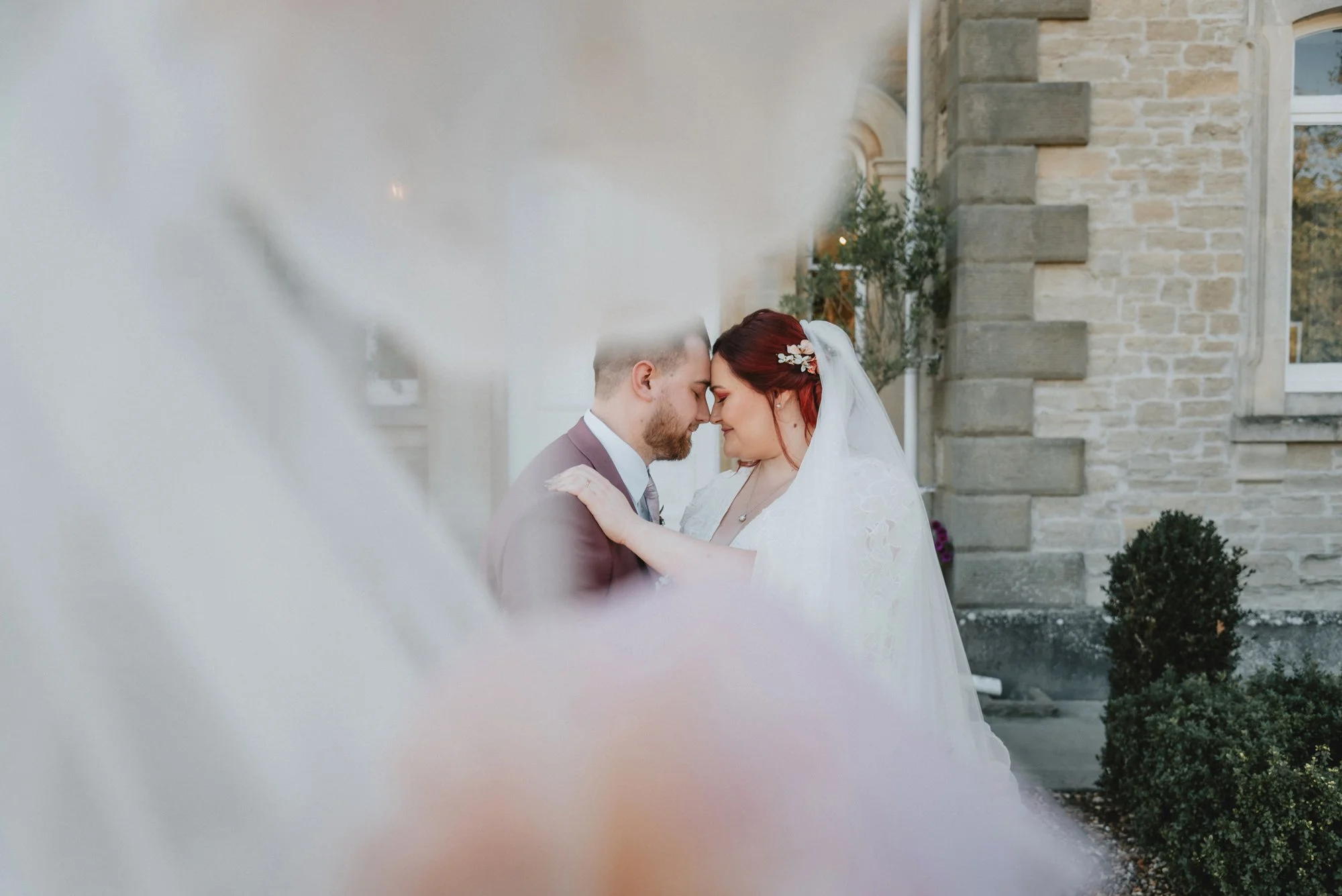 A bride and groom with foreheads touching, embracing outdoors in front of a stone building, during wedding photos.