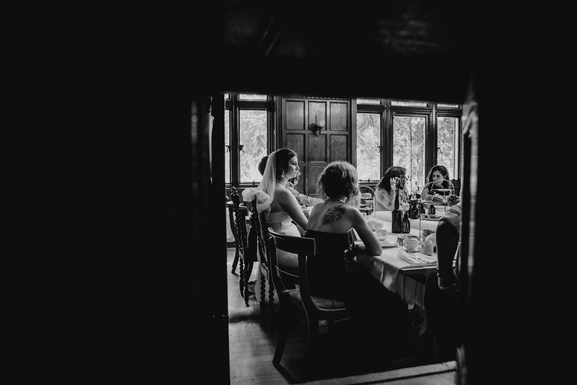 Women having tea or coffee at a decorative table in a vintage-style room with large windows, viewed through a small opening.