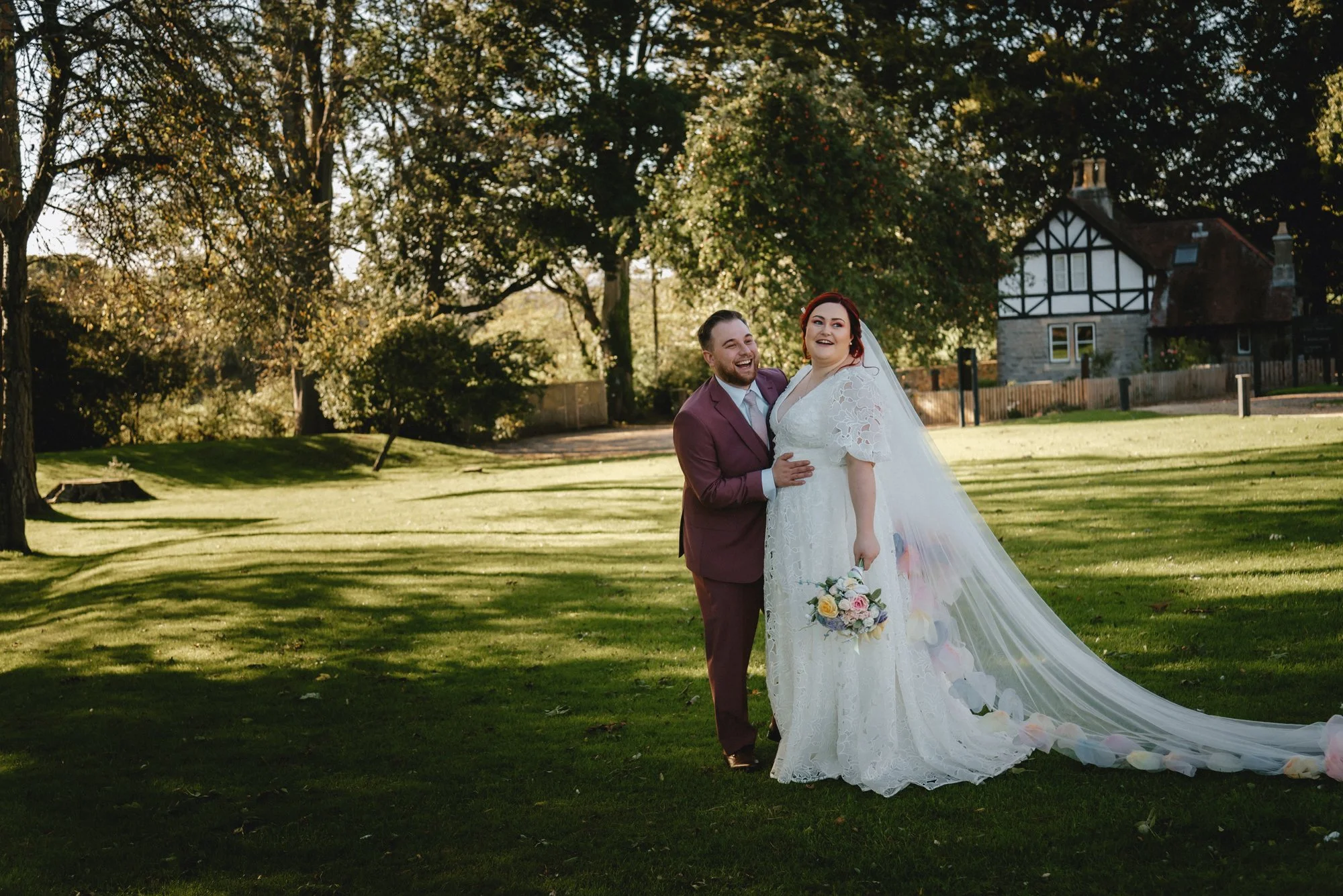 A newly married couple stands on a grassy lawn, with the bride holding a bouquet and wearing a wedding dress with a long veil, while the groom wears a maroon suit. They are smiling and hugging, with trees and a house in the background on a sunny day.