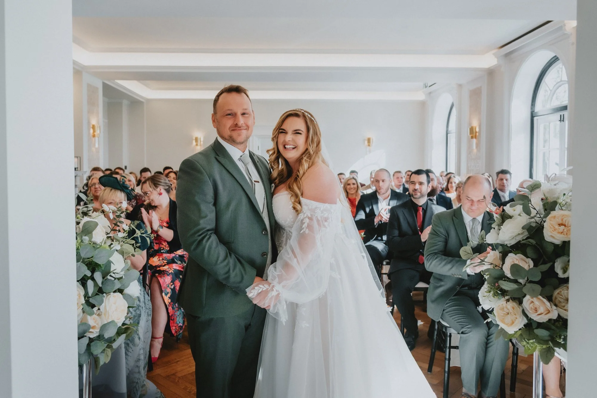 Bride and groom smiling at a wedding ceremony, surrounded by seated guests in a bright, elegantly decorated room with large windows and floral arrangements.