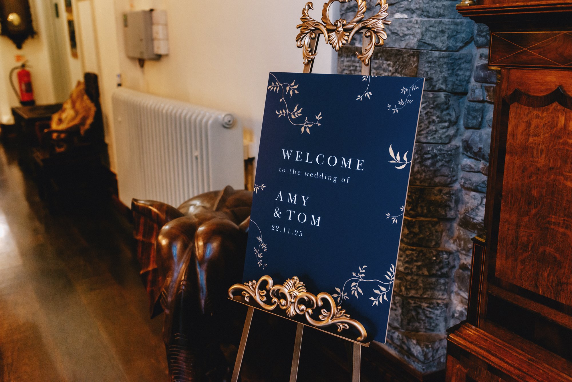 Wedding welcome sign decorated with floral designs, indicating the marriage of Amy and Tom on November 25th, placed inside a warmly lit venue near a leather sofa and brick wall.