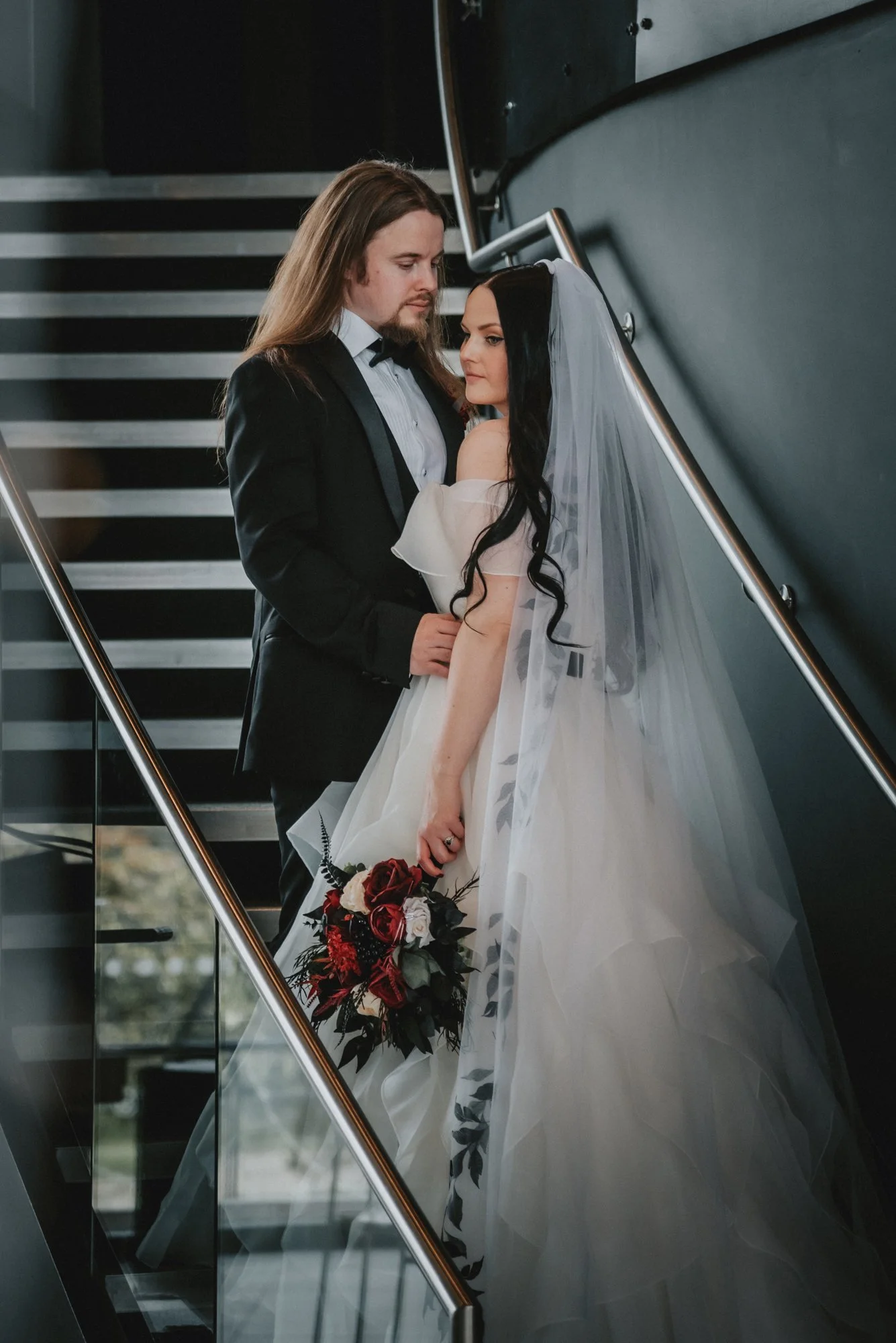 A bride and groom standing on a staircase, gazing at each other, with the bride holding a bouquet of red and white flowers.
